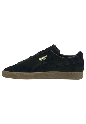 Sorte ruskinds sneakers med en flad, brun gummisål. Egenskaber omfatter runde snørebånd og et guldlogo på siden. Klassisk lav-top design.