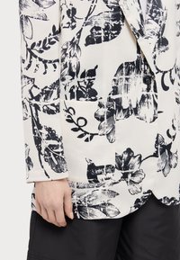 Hvid blazer med sort blomsterprint, der har en reverskrave, en enkelt knaplukning og sidelommer, samt en glat stofstruktur.