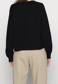 Pull en tricot noir avec une coupe oversize, poignets et ourlet côtelés, doté d'un col rond et d'une finition texturée. Associé à un pantalon beige.