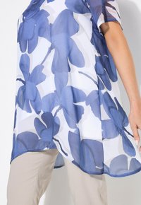 Leichte, durchsichtige Bluse mit großen blauen Blumenmustern auf weißem Hintergrund. Abgerundeter Saum, kurze Ärmel, lässige Passform.