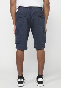 Férfi tengerészkék cargo sortban, amely több zsebből áll, fehér pólóban, fekete zokniban és fekete-fehér sportcipőben áll egy fehér háttér előtt.