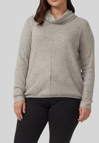 Frau, die einen beigen Waffelpullover mit langen Ärmeln und Trichterkragen sowie schwarze Hosen vor einem einfarbigen grauen Hintergrund trägt.