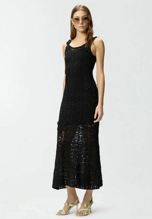 Vrouw draagt een mouwloze zwarte kanten maxi-jurk met bloemmotieven, beige sandalen met hoge hakken en oversized ronde zonnebril, staand.