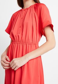 Robe corail à manches courtes, col froncé et taille élastique. Présente des détails de boutons à l'avant, avec une coupe légèrement ample.