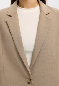 Beige kavaj med snedslåda, enkel knappstängning och strukturerad tyg, bärs över en vit topp. Närbild av krageområdet.