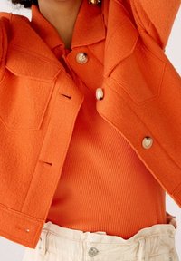 Orange ulljacka med texturerad yta, med två framfickor och stora knappar, lager på en ribbad orange topp och en krämfärgad midjeband.