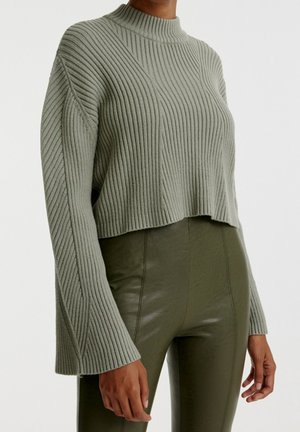 Femme portant un pull court en tricot côtelé olive clair avec des manches évasées et un pantalon ajusté taille haute de couleur olive foncé.