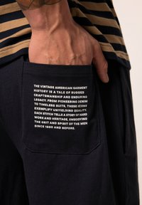 Pantalones cortos de marinero con una tela texturizada presentan un bolsillo izquierdo que muestra texto en blanco sobre la historia de la confección estadounidense y la artesanía.