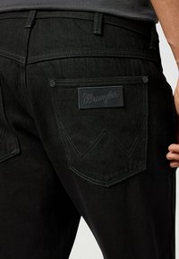 Jeans negri din denim, având un buzunar posterior cu un model distinctiv de cusătura "W" și o etichetă rectangulară din piele embosată cu „Wrangler”. 
