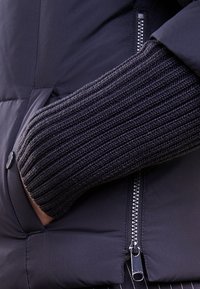 Chaqueta acolchada negra con una manga de punto acanalado oscuro en contraste visible. Presenta un cierre de cremallera y un acento de botón redondo.