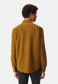 Camisa de color mostaza con mangas largas, puños abotonados y cuello clásico. Textura suave y detalle en el dobladillo curvado.