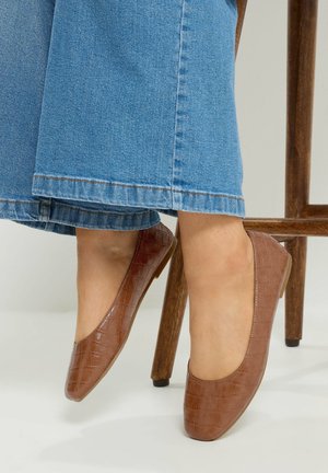 Voeten met bruine, getextureerde ballerina's, gecombineerd met wijd uitlopende blauwe spijkerbroeken, rustend nabij een houten stoelbeen.