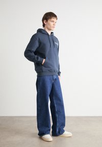 Marineblå hettegenser med kengurulomme og justerbare snorer, kombinert med vide blå jeans og beige sneakers.