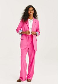 Różowy garnitur skrojony na miarę z dwurzędową marynarką i szerokimi spodniami. Wylot rękawów odsłania wzorzystą tkaninę. W zestawieniu z butami w panterkę.