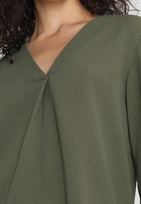 Blusa verde oliva con scollatura a V, caratterizzata da una texture sottile e una parte anteriore drappeggiata. Tessuto leggero che scivola delicatamente sul corpo.