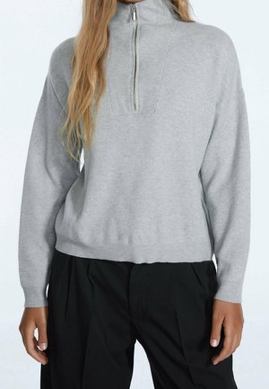 Vrouw draagt een lichtgrijze trui met lange mouwen en een voorrits, gecombineerd met zwarte, loszittende broek met hoge taille, staand met ontspannen handen.