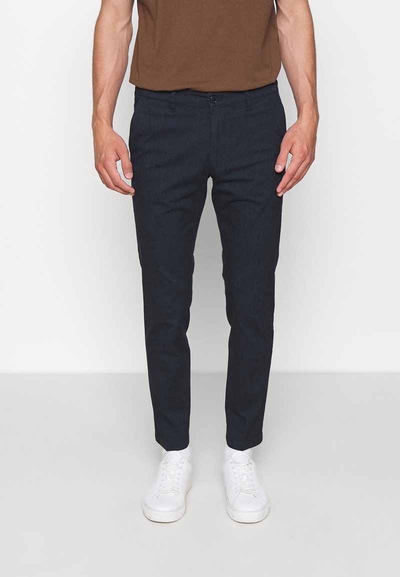 Pantalons slim-fit bleu marine en tissu texturé, dotés de poches avant et d'une fermeture à bouton, associés à des baskets blanches.