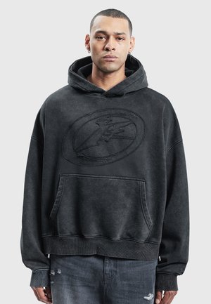 Hombre que lleva una sudadera negra de gran tamaño con un gran logo en relieve en el pecho y vaqueros oscuros desgarrados, de pie frente a un fondo liso.