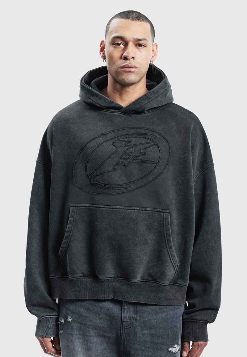 Homme portant un sweat à capuche noir oversized avec un grand logo embossé sur la poitrine et un jean déchiré foncé, debout devant un fond uni.