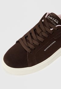 Brown suede sneaker featuring round toe, flat white rubber sole, and matching brown laces. “Scotch & Soda” logo printed on the side.