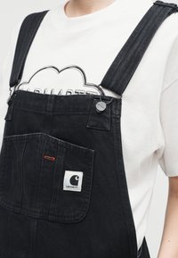 Άτομο που φοράει μαύρη τζιν φόρμα με λογότυπο στην μπροστινή τσέπη πάνω από λευκό μπλουζάκι με μαύρο περίγραμμα γραφικού.