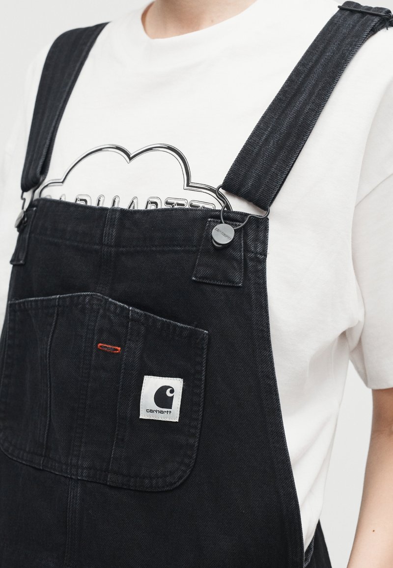 Person trägt schwarze Denim-Latzhose mit Logo auf der Vorderseite über einem weißen T-Shirt mit einem schwarzen konturierten Grafikdruck.