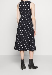 Žena v černých midi šatech bez rukávů s bílými puntíky a černými kotníkovými botami, stojící na bílém pozadí.