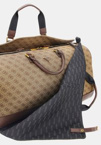 Sac de voyage brun à motifs avec des accents en cuir, matériel doré et doublure intérieure noire présentant un imprimé de logo répété. Sangle réglable visible.