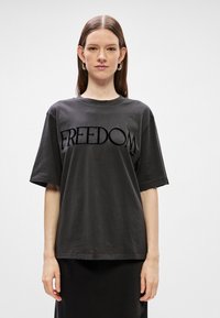 Μαύρη βαμβακερή μπλούζα με κοντά μανίκια, που φέρει τη λέξη "ΕΛΕΥΘΕΡΙΑ" σε ανυψωμένη, βελούδινη υφή στην περιοχή του στήθους.