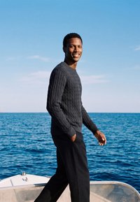 Homme portant un pull sombre et un pantalon, souriant, debout sur un bateau avec l’océan bleu et un ciel clair en arrière-plan.
