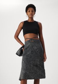 Fekete bordázott crop top, szürke midi szoknyával, amely texturált felülettel és fonott öv részlettel rendelkezik. A modellt egy fekete táska tartása jellemzi.