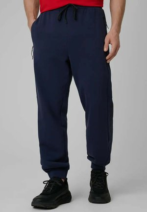 Mężczyzna ubrany w granatowe spodnie dresowe z elastycznym pasem i czarne trampki, stojący na jednolitym szarym tle.