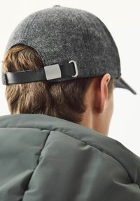 Grå tekstureret kasket med en sort læderrem og metalspænde, set bagfra mod en grøn polstret jakke.