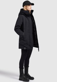 Svart vattentålig parka med hög krage, dragkedja och tryckknappar, sidofickor, tillsammans med svarta leggings och kraftiga svarta stövlar.