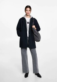 Marinblårandig kappa med klippt krage, bärs över en grå hoodie och vit t-shirt, kombinerat med grå vida byxor och svarta ankelstövlar.