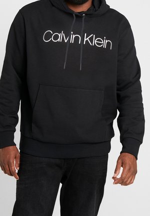 Dorosły w czarnej bluzie z kapturem Calvin Klein z przednią kieszenią i czarnych spodniach, stojący na jednolitym jasnym tle.
