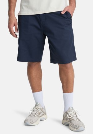 Mężczyzna w granatowych szortach, białych skarpetkach sportowych i beżowych butach sportowych, stojący z rękami na biodrach na białym tle.