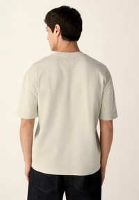 Camiseta de manga corta gris claro, hecha de un tejido texturizado con un cuello redondo. Presenta un diseño minimalista y un corte ligeramente holgado.