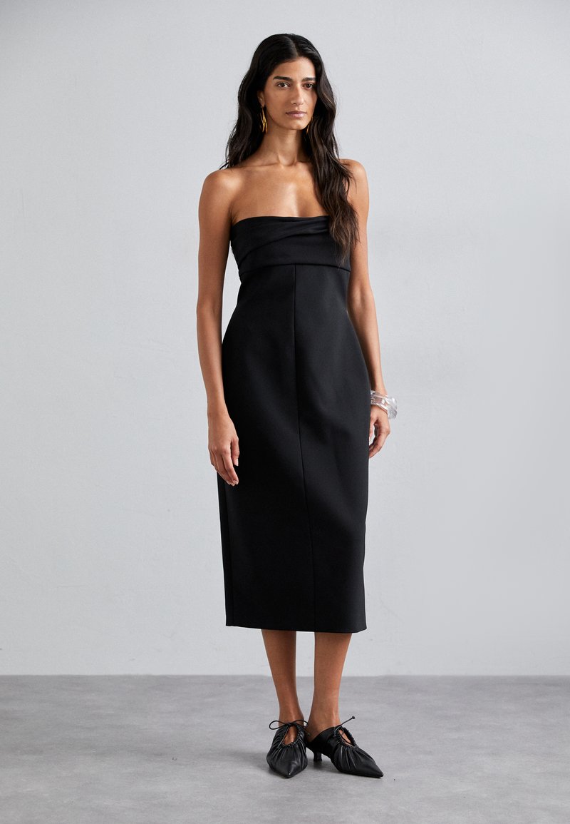 Schwarzes, trägerloses Midi-Kleid mit tailliertem Oberteil und fließendem Rock. Glatte Stoffstruktur, keine sichtbaren Muster, kombiniert mit schwarzen Schuhen.
