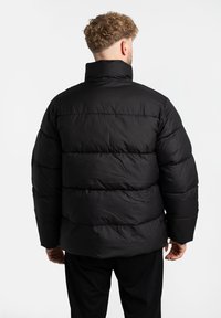 Zwarte pufferjack met een hoge kraag, zachte textuur, horizontale quilt en een ruime pasvorm. Zichtbare stiksels en geen zichtbare hardware.