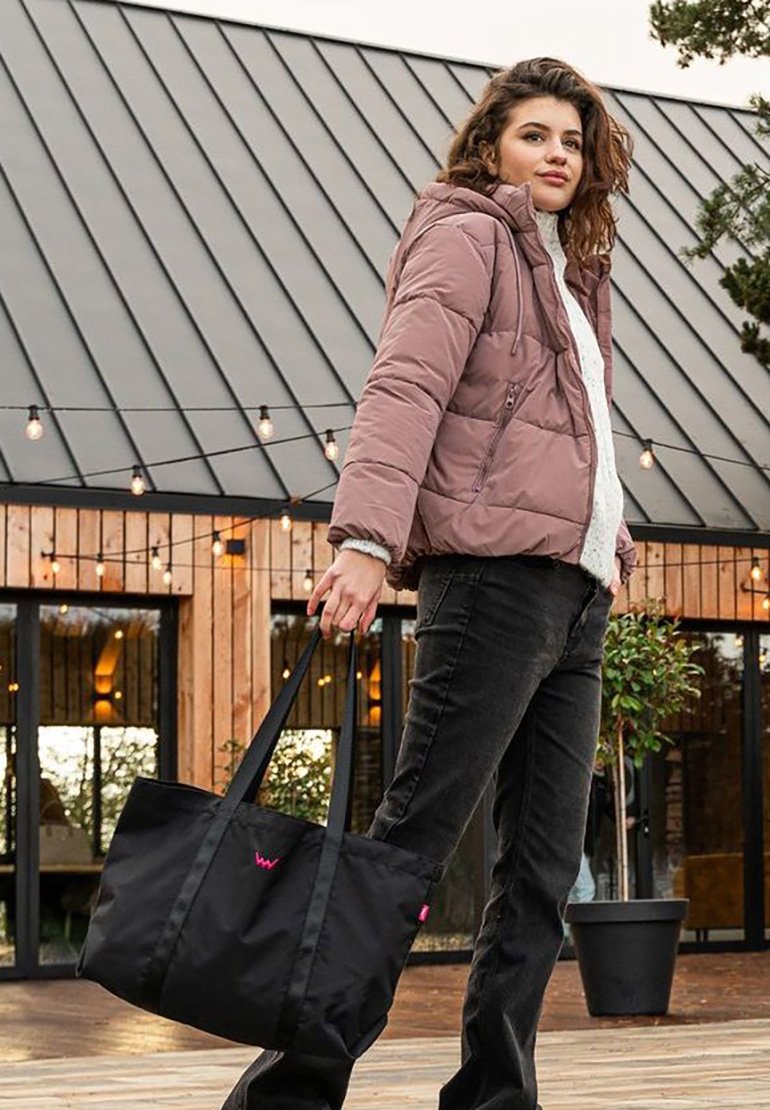 Black tote bag with pink branding, made of durable fabric, featuring two long handles. Worn with a light pink puffer jacket and black pants.