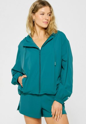 Frau mit blonden Haaren, die eine tealfarbene Kapuzenjacke mit Reißverschluss und passende Shorts trägt, schaut zur Seite und hat eine Hand in der Tasche.