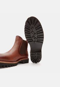 Ruskea nahkainen Chelsea-saapas, jossa on joustava sivupaneeli ja syvällä kuvioitu kumipohja pitävyyttä varten. Vahvistetut saumat reunalla.