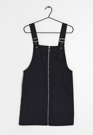 Czarna sukienka na szelkach w cienkie prążki, z przednim suwakiem, regulowanymi paskami z klamrami oraz dwoma kieszeniami z przodu, zawieszona na czarnym wieszaku.