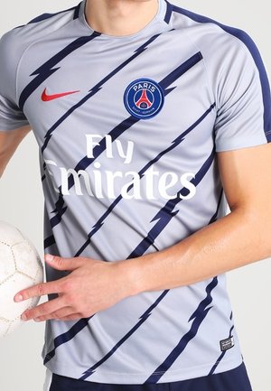 Osoba ubrana w szary strój Paris Saint-Germain z granatowymi błyskawicami, trzyma białą piłkę nożną, pokazując tułów i ramiona.