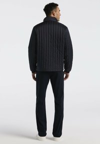 Man in een zwart gewatteerd jack, zwarte broeken en witte sneakers, staand met zijn rug naar de camera tegen een plain witte achtergrond.