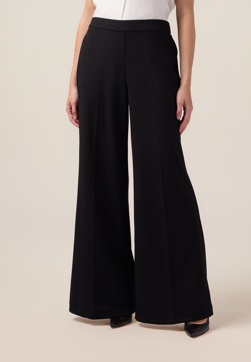 Pantalon noir à jambes larges en tissu lisse, avec une taille haute et une coupe ajustée qui descend jusqu'aux chevilles.
