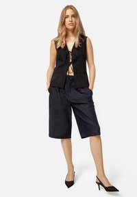 Vesta neagră, fără mâneci, cu un nod frontal, asociată cu pantaloni scurți largi din denim închis și pantofi cu vârf ascuțit negri. Design simplu, contemporan.