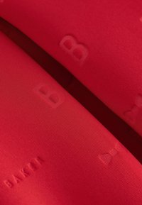 Červená tkanina s raženým nápisem a vzory, na které je uvedeno slovo "BAKER" a mašle. Textura je hladká a mírně lesklá.
