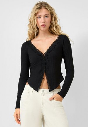 Blonde vrouw met golvend haar, gekleed in een zwart V-hals top met lange mouwen en kanten afwerking, gecombineerd met gebroken witte broek, één hand in de zak.